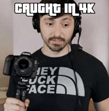 a man wearing headphones and a shirt that says hey fuck face holds a camera