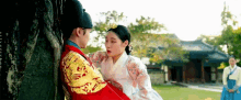 a man and a woman are leaning against a tree . the woman is wearing a traditional korean dress .