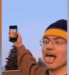 a man wearing glasses and a yellow beanie holds up a cell phone