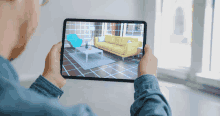 a man is holding a tablet that shows a living room with a yellow couch and blue chair