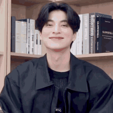 a young man in a black jacket is smiling in front of a bookshelf filled with books including one titled luxury hotels