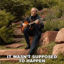 a man sits on a rock playing a guitar with the words it wasn 't supposed to happen behind him