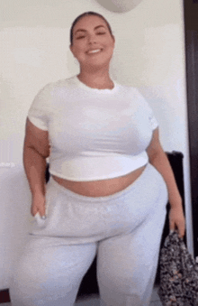 a woman is standing in a room wearing a white crop top and gray sweatpants .