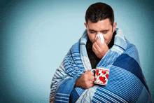 a man wrapped in a blanket is blowing his nose while holding a cup