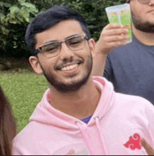 a man with a beard wearing glasses and a pink hoodie is smiling while holding a drink .