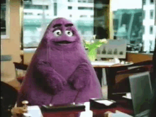 a purple stuffed animal is standing in front of a desk in an office .