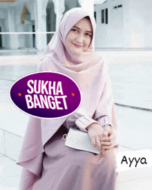 a woman wearing a pink hijab sits in front of a purple sign that says sukha banget