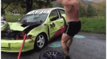 a man without a shirt is standing in front of a damaged car with a sign that says fuel on it .