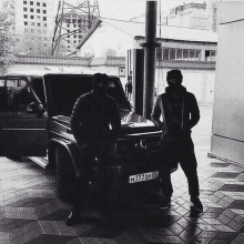 a black and white photo of two men standing next to a car with a license plate that says m777 op 05