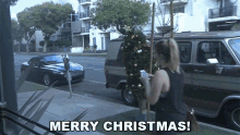 a woman is holding a christmas tree in front of a van and says merry christmas