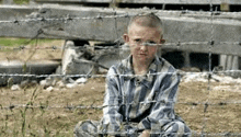 a young boy in a striped shirt is sitting behind barbed wire fence .