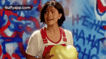 a woman in overalls is holding a heart shaped balloon in front of a graffiti wall .