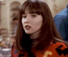 a woman with long brown hair and bangs is wearing a red and black sweater and looking at the camera .