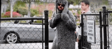 a man in a fur coat is standing in front of a chain link fence with a sign that says please close gate behind you