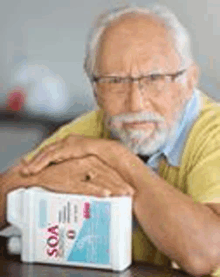 an older man with a beard and glasses is sitting at a table with a bottle of soa .