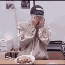 a man wearing a baseball cap is covering his face while sitting at a table with a bowl of food and chopsticks .