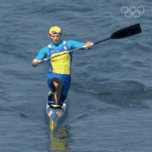 a man in a blue and yellow outfit is paddling a canoe in the water with the olympic rings in the background