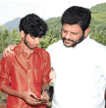 a man in a white shirt stands next to another man in a red shirt looking at a cell phone