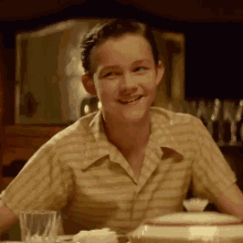 a young man in a striped shirt is smiling while sitting at a table with a glass of water .