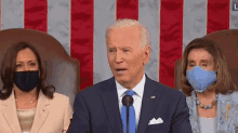 a man in a suit and tie is speaking into a microphone in front of two women wearing masks .