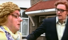 two men are standing next to each other in front of a house . one of the men is wearing glasses and a hat .