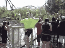 a woman in a neon green shirt is standing on a fence in front of a roller coaster at an amusement park .