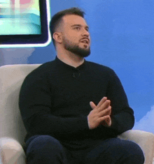 a man with a beard is sitting in a chair with his hands folded in front of him