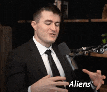 a man in a suit and tie is talking into a microphone with the word aliens visible in the corner