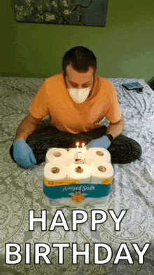 a man wearing a mask and gloves is sitting on a bed with a birthday cake made out of toilet paper rolls .