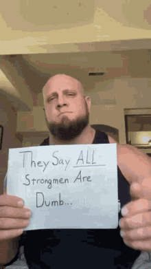 a bald man with a beard is holding up a sign that says they say all strongmen are dumb