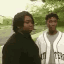 two men are standing next to each other in a park . one of the men is wearing a baseball jersey .