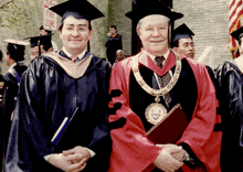 a man in a graduation cap and gown stands next to another man in front of a fire zone sign