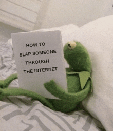 a kermit frog laying on a bed holding a card that says " how to slap someone through the internet "