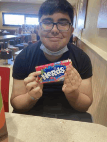 a young man wearing glasses is sitting at a table holding a bar of nerds candy .