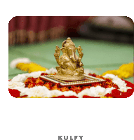 a statue of ganesha sits on a table with flowers and the word kulfy below it