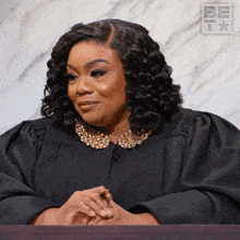 a woman in a black gown with a gold collar sits at a table in front of a sign that says be