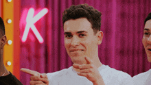 a man in a white shirt is pointing at a pink curtain with the letter k in the background