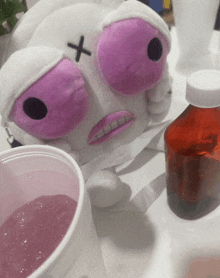 a stuffed animal with a cross on its face sits next to a cup of liquid and a bottle of liquid