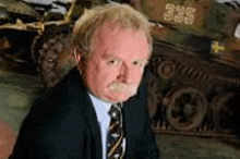 a man in a suit and tie is sitting in front of a military vehicle .