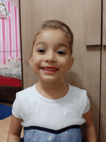 a little girl wearing a white shirt and a blue dress smiles in front of a mirror