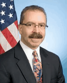 a man with a mustache is wearing a suit and tie and standing in front of an american flag .