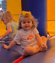 a little girl wearing a rainbow shirt is sitting on a trampoline