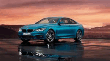 a blue bmw 4 series coupe is parked on a wet road in front of a sunset .