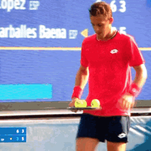 a man in a red shirt holds a tray of tennis balls in front of a scoreboard that says 63