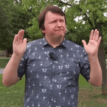 a man in a blue shirt with palm trees on it is standing in a park with his hands in the air .