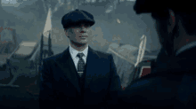 a man in a suit and tie is talking to another man in a flat cap .