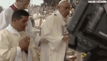 a man in a white robe is praying in front of a camera while a group of priests stand behind him .