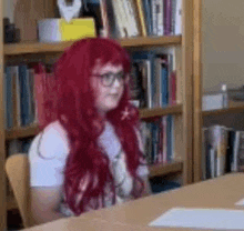 a girl with red hair and glasses is sitting at a table in front of a bookshelf in a library .