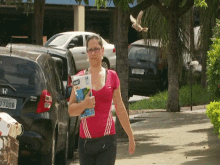 a woman in a pink shirt is walking down the street with a magazine in her hand and a honda car behind her