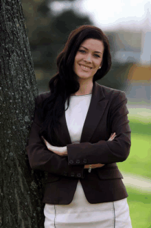 a woman leans against a tree with her arms crossed and smiles